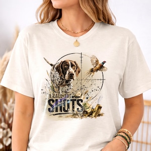 May include: Cream-colored t-shirt featuring a hunting-themed graphic. The design includes a dog, a pheasant, and a scope, with the text "I CALL THE SHOTS" in a stylized font. Two spent shotgun shells are also visible.