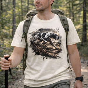 May include: Cream-colored t-shirt featuring a black and white dog illustration in a dynamic pose. The graphic design includes splatters and a natural landscape. The person is wearing a backpack and holding a hiking stick.