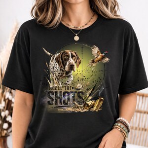 Puede incluir: Camiseta negra con un diseño de perro de caza, un faisán y el texto "CALL THE SHOTS". El diseño incluye una mira telescópica, hierba y dos casquillos dorados.