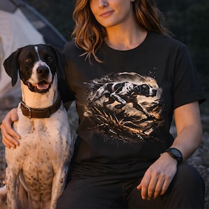 May include: Black t-shirt with a graphic of a Pointer dog in action. The dog is black and white, set against a natural backdrop. A real Pointer dog with a brown collar sits next to the person wearing the shirt.