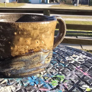 May include: A handmade ceramic mug with a textured, brown exterior and a blue-grey base. The mug has a rounded shape with a handle and is sitting on a black surface.