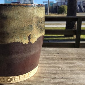 May include: A handmade ceramic coffee mug with a two-tone glaze. The top half is a muted green and tan, transitioning to a deep brown. The word "COFFEE" is stamped on the base. The mug sits on a wooden surface.