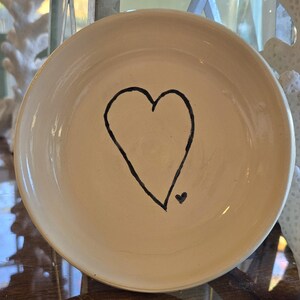 May include: A white ceramic dish with a black hand-drawn heart design. The heart has a small heart at the bottom.