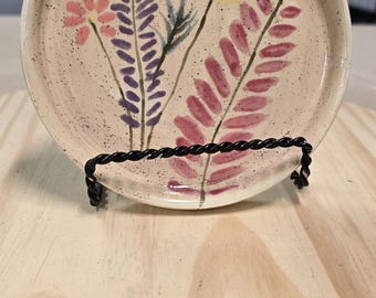 May include: A small, round, ceramic dish with a speckled beige glaze. The dish features a hand-painted floral design with pink, purple, and yellow flowers.