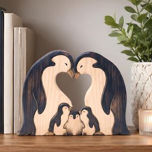 Könnte beinhalten: Eine Holzpuzzle-Skulptur mit zwei großen erwachsenen Pinguinen und drei kleinen Pinguinküken. Die erwachsenen Pinguine stehen einander gegenüber und bilden einen herzförmigen Freiraum zwischen ihren Hälsen. Das Holz hat natürliche helle und dunkel gebeizte Oberflächen.