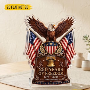 Op de afbeelding: Een helder acryl herdenkingsstuk met een adelaar met uitgespreide vleugels, Amerikaanse vlaggen en de tekst "250 YEARS OF FREEDOM 1776-2026". Het ontwerp bevat een bel en sterren, ter viering van de 250e verjaardag.