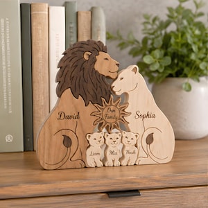 Può includere: Un puzzle in legno raffigurante una famiglia di leoni con un leone marrone scuro, una leonessa color sabbia chiaro e tre cuccioli. Il pezzo include i nomi incisi David, Sophia, Liam, Mia e Noah, con un emblema solare centrale con la scritta Our Family.