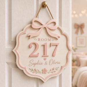 Op de afbeelding: Een crèmekleurig houten deurbordje met een lichtroze rand en een bijpassende roze strik aan de bovenkant. Het bordje bevat de tekst "ROOM 217" in grote roze cijfers, gevolgd door de namen "Sophia & Olivia" en een klein bloemmotief onderaan.