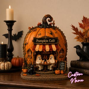 Può includere: Una struttura decorativa a forma di zucca etichettata Pumpkin Café con una tenda a righe, che mostra due figure di fantasmi sedute a un tavolo. L'esterno è decorato con foglie autunnali, un gatto nero e un cartello Coffee Cookies Cozy Vibes.