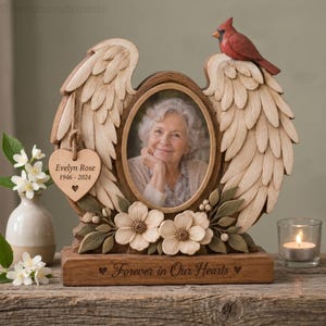 May include: A wooden memorial photo frame featuring carved angel wings, a red cardinal bird on top, and a heart-shaped tag inscribed with Evelyn Rose 1946-2024. The base is engraved with the text Forever in Our Hearts and floral accents.