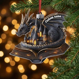 Peut inclure: Une décoration suspendue plate représentant un dragon sombre et texturé enroulé autour d'un château détaillé posé sur un livre ouvert. L'aile du dragon porte l'inscription 'Custom name' en blanc, et un ruban rouge est fixé au sommet.