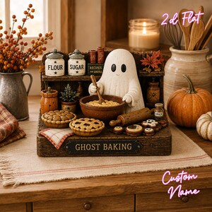 May include: A decorative tabletop display featuring a white ghost figurine baking in a kitchen setting. The piece includes miniature jars labeled Flour and Sugar, a bowl, a rolling pin, two pies, and a sign that reads GHOST BAKING in black text.