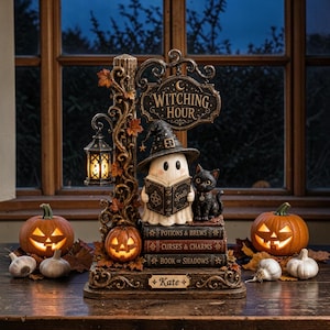 Peut inclure: Une figurine décorative d'Halloween représentant un fantôme avec un chapeau de sorcière lisant un grimoire, un chat noir et une lanterne sur un poteau. La base porte l'inscription "Witching Hour" et une plaque au nom de "Kate".