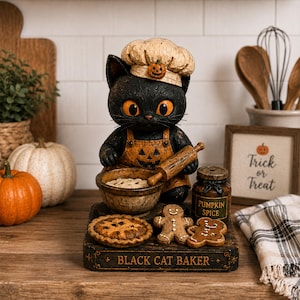 Puede incluir: Una figura de gato negro vestido de panadero, con un gorro de chef con un detalle de calabaza y un delantal naranja con una cara de calabaza. El gato sostiene un rodillo sobre un cuenco, con un pastel, dos galletas de jengibre y un tarro etiquetado Pumpkin Spice sobre una base marcada Black Cat Baker.
