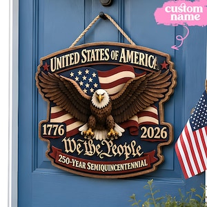 May include: A wooden sign with the text "UNITED STATES OF AMERICA" and "We the People". It features an eagle, an American flag, and the years 1776 and 2026. The sign is designed for the 250-Year Semiquincentennial.