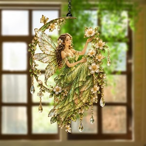 May include: A decorative hanging ornament featuring a fairy with translucent wings sitting on a crescent moon. The fairy wears a flowing green dress and is surrounded by white flowers, green leaves, and dangling clear crystal teardrop accents.