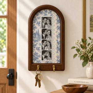 Può includere: Un portachiavi da parete in legno ad arco con cornice marrone scuro, sfondo con motivo floreale blu e bianco, una striscia verticale di pizzo color crema e una striscia fotografica di una coppia. Due ganci color ottone reggono delle chiavi.