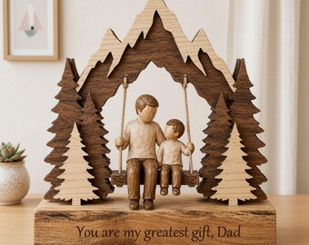 May include: A wooden sculpture featuring a father and child on a swing, set against a mountain and forest backdrop. The base reads "You are my greatest gift, Dad." The sculpture is made of light and dark brown wood.