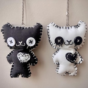 May include: Two plush cat-shaped ornaments hanging from a wooden rod by silver ball chains. One is black with white button eyes and a white heart patch, the other is white with black button eyes and a black heart patch, both featuring visible white stitching.
