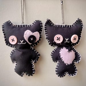 May include: Two black fabric plush cat ornaments with white whip-stitched edges and silver ball chains. One cat features a pink heart patch over one eye with a black button, while the other has pink button eyes and a pink heart patch on its chest.