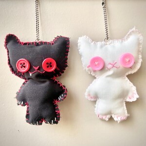 May include: Two plush cat-shaped keychains hanging from a wooden rod. One is black with red stitching and red button eyes, the other is white with pink stitching and pink button eyes. Both feature metal ball chains for attachment.