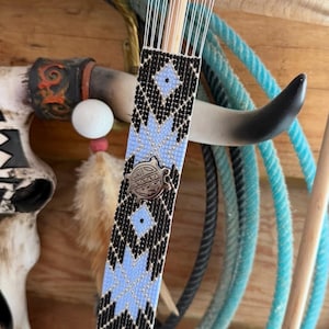 May include: A beaded bookmark with a geometric pattern in black and light blue, featuring a silver-toned turtle charm. The bookmark is attached to a wooden weaving tool, with a wooden background and a blue rope.