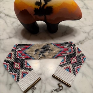 Puede incluir: Una pulsera de cuentas con un diseño de caballo y una figura de oso de cerámica. La pulsera presenta un motivo central de caballo, enmarcado por patrones geométricos rojos, negros y azules. La figura del oso tiene un diseño de paisaje desértico.