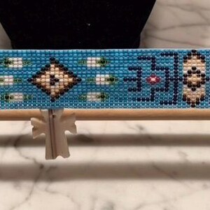 May include: A beaded turquoise bracelet with a geometric pattern is displayed on a wooden loom. The bracelet features a central diamond design flanked by symmetrical shapes in white, black, and pink. The loom is made of light-colored wood.