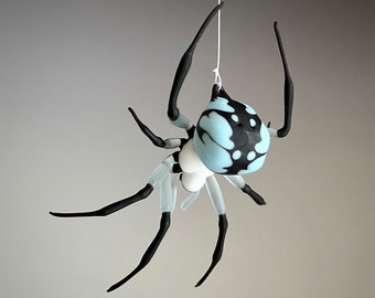 May include: A glass spider sculpture hanging from a white string. The spider is black and white with a blue and black patterned abdomen.