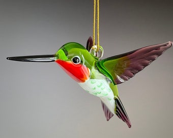 May include: A glass hummingbird ornament with a green body, red throat, white and green markings, and purple wings. The ornament is hanging from a gold string.