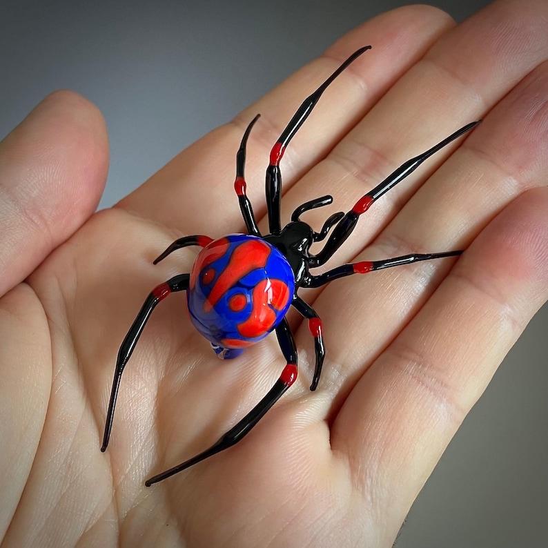 May include: A glass spider sculpture with a black body and red and blue patterned abdomen. The spider is posed with its legs outstretched.