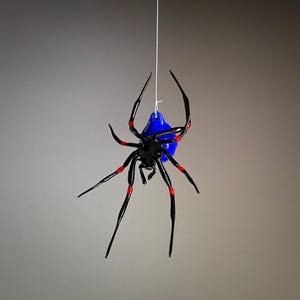 May include: A black glass spider with red accents hanging from a white string. The spider has a blue glass body.