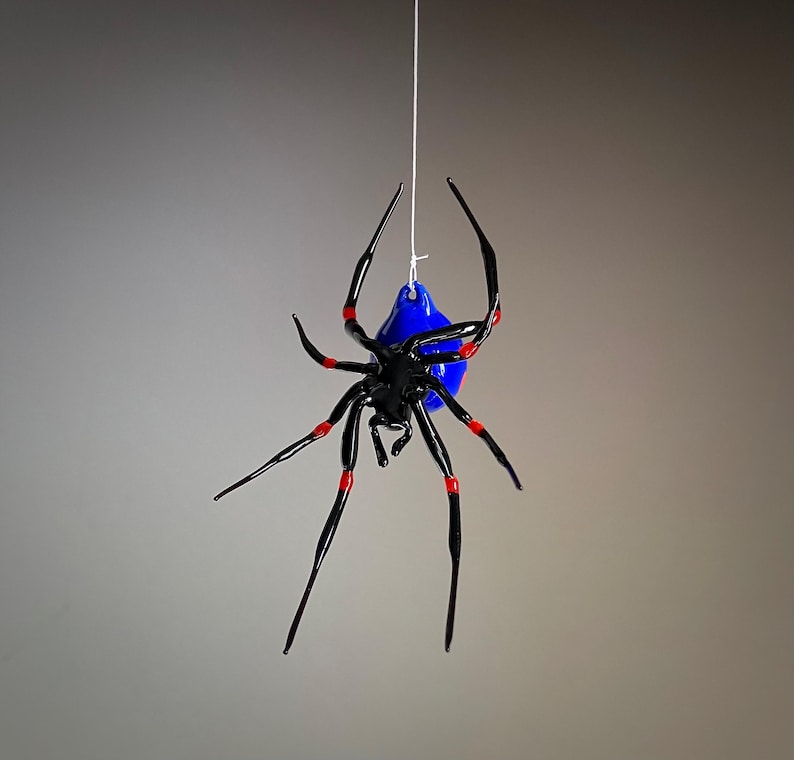 May include: A black glass spider with red accents hanging from a white string. The spider has a blue glass body.
