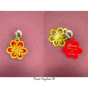 Puede incluir: Llaveros con forma de flor en rojo, amarillo y plateado. Un llavero tiene el texto "Bloom where you are planted". Los llaveros están sobre un fondo rosa. Los llaveros están sujetos a un llavero plateado.