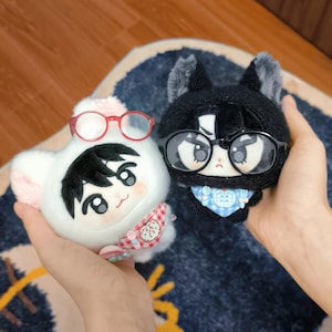 Puede incluir: Dos muñecos de peluche, uno blanco y otro negro, se sostienen en las manos. El muñeco blanco tiene detalles rosas, gafas rojas y una bufanda estampada. El muñeco negro tiene gafas negras y una bufanda azul estampada. Ambos muñecos tienen caras de dibujos animados.
