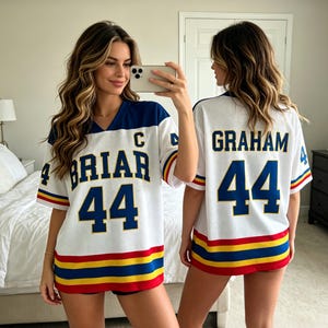 Op de afbeelding: Wit shirt met blauwe, rode en gele strepen. Het shirt heeft de naam "BRIAR" en het nummer "44" op de voorkant. De achterkant van het shirt heeft de naam "GRAHAM" en het nummer "44". Het shirt heeft een blauwe kraag.