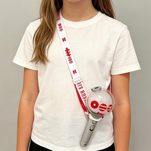 May include: A white strap with red text and a gray and clear light stick. The strap says "BTS ARMY" and has red symbols. The light stick has a clear globe with red accents and a gray handle.