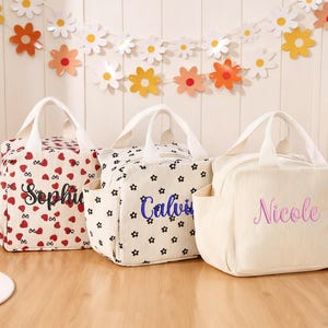 Op de afbeelding: Drie gepersonaliseerde draagtassen met witte handvatten en zijvakken. Eén tas is crèmekleurig met rode hartjes, een andere is wit met zwarte sterren en de derde is crèmekleurig met verticale lijnen. Elke tas heeft een geborduurde naam.