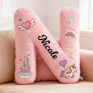 Puede incluir: Cojín rosa de felpa con la letra "N" y el nombre "Nicole" en escritura negra. Decorado con gráficos de dibujos animados que incluyen un corazón alado, un castillo, un arcoíris, un unicornio y una estrella fugaz. Un cojín decorativo para la habitación de un niño.