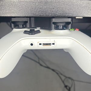 Könnte beinhalten: Ein weißer Xbox-Controller ist mit einem schwarzen Plastikhalter an einem Schreibtisch befestigt. Der Controller zeigt nach unten, die Tasten und Joysticks sind sichtbar.