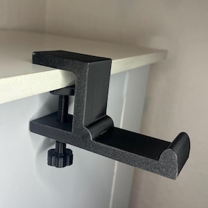 Könnte beinhalten: Schwarzer Kopfhörerständer, an einer weißen Oberfläche befestigt. Der Ständer hat ein C-förmiges Design mit einer gepolsterten Auflage für die Kopfhörer und einer Schraubklemme zur Befestigung. Der Ständer besteht aus mattem, schwarzem Material.
