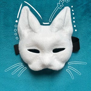 Peut inclure: Un masque de chat en feutrine blanche avec des oreilles pointues et des ouvertures noires pour les yeux. Le masque comporte une sangle élastique noire de chaque côté. Des dessins au trait blanc représentant des moustaches et des détails d'oreilles sont superposés autour du masque sur le fond sarcelle.