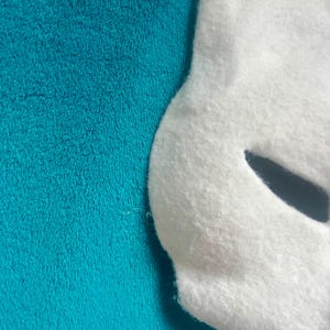 May include: A white felt mask with a curved edge and a single dark, almond-shaped eye cutout. The mask has a soft, fuzzy texture and is positioned against a solid, vibrant teal background with a plush, fleece-like appearance and texture.