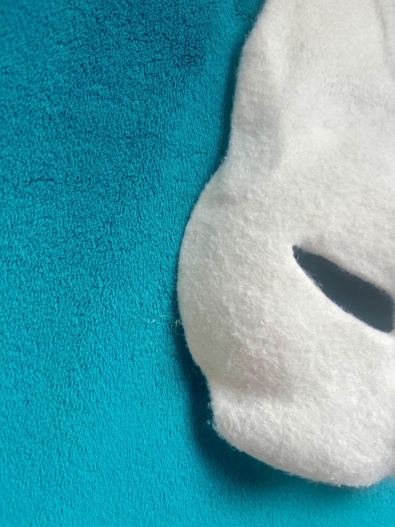 May include: A white felt mask with a curved edge and a single dark, almond-shaped eye cutout. The mask has a soft, fuzzy texture and is positioned against a solid, vibrant teal background with a plush, fleece-like appearance and texture.