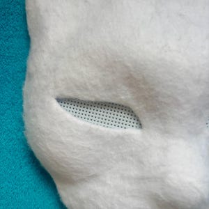 May include: A white, fuzzy, animal-shaped mask featuring a smooth, felt-like texture. The eye opening is almond-shaped and covered with a white, fine-mesh screen material that allows for visibility while maintaining the mask's overall structure.