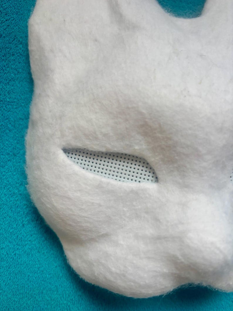 May include: A white, fuzzy, animal-shaped mask featuring a smooth, felt-like texture. The eye opening is almond-shaped and covered with a white, fine-mesh screen material that allows for visibility while maintaining the mask's overall structure.