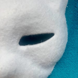 May include: A white, felted mask featuring a smooth, rounded surface and a single, elongated, horizontal eye opening. The material has a soft, fuzzy texture throughout, with clean, curved edges defining the shape of the facial structure.