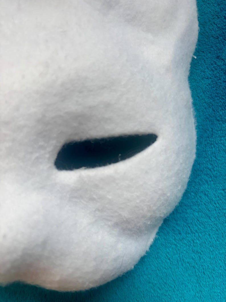 May include: A white, felted mask featuring a smooth, rounded surface and a single, elongated, horizontal eye opening. The material has a soft, fuzzy texture throughout, with clean, curved edges defining the shape of the facial structure.