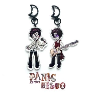 May include: Two acrylic keychains featuring cartoon illustrations of a singer and a guitarist. The singer holds a microphone, and the guitarist plays a cream-colored guitar. Both figures have dark hair and are dressed in black and white outfits. The text "Panic at the Disco" is at the bottom.