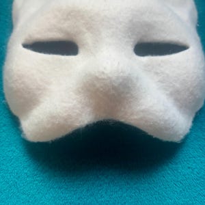 May include: A white felt mask featuring a stylized animal shape with pointed ears and two horizontal, almond-shaped eye openings. The surface has a soft, fuzzy texture, and the mask is molded to follow the contours of a face with a defined nose bridge.